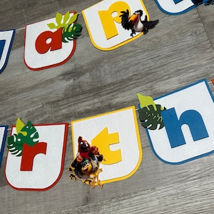 May include: A colorful banner with the letters "Rio" and a toucan and macaw bird illustration. The banner is made of cardstock and has a white background with a textured pattern. The letters are in different colors, including red, yellow, blue, and green.