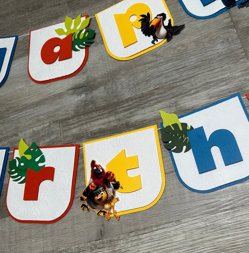 May include: A colorful banner with the letters "Rio" and a toucan and macaw bird illustration. The banner is made of cardstock and has a white background with a textured pattern. The letters are in different colors, including red, yellow, blue, and green.