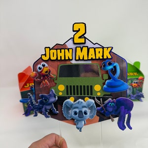 Pode incluir: Um topper de bolo de jipe verde com uma cobra azul, uma aranha roxa, um coala cinza, um dragão azul e cinza e um caranguejo vermelho e laranja. O topper também inclui o número 2 e o nome "JOHN MARK".