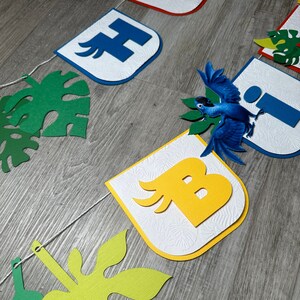 May include: A colorful banner with the letters "H", "I", "B" and a blue bird with green leaves. The letters are on white backgrounds with a textured pattern. The banner is strung together with white string.