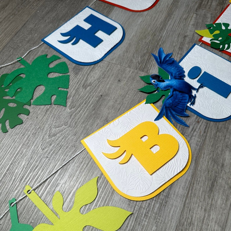 May include: A colorful banner with the letters "H", "I", "B" and a blue bird with green leaves. The letters are on white backgrounds with a textured pattern. The banner is strung together with white string.