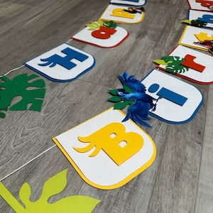 May include: A colorful felt banner with the letters "Happy Birthday" in white, blue, yellow, and red. The letters are decorated with green leaves and a blue bird.