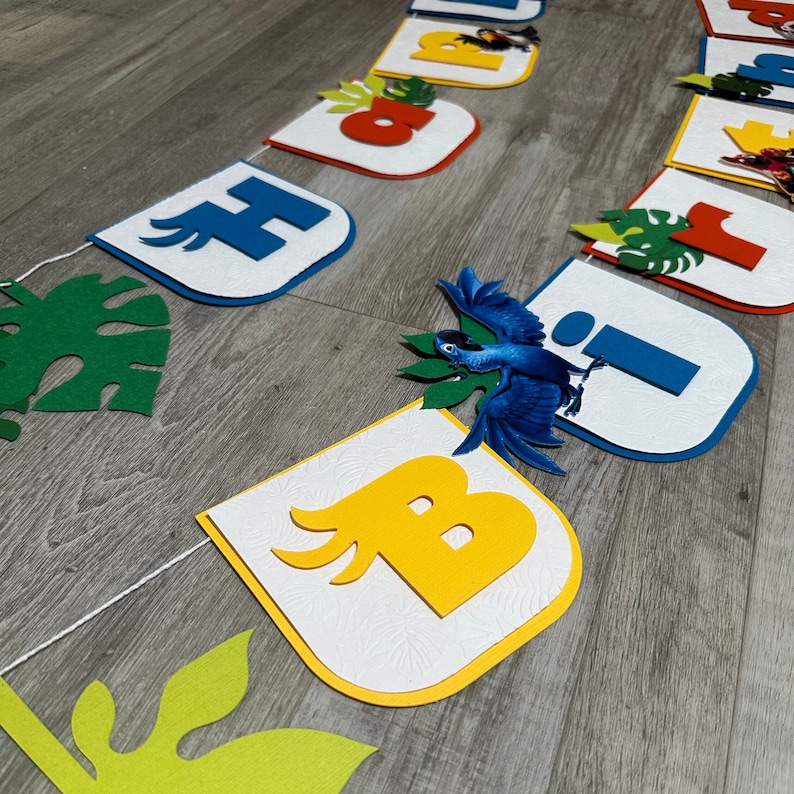 May include: A colorful felt banner with the letters "Happy Birthday" in white, blue, yellow, and red. The letters are decorated with green leaves and a blue bird.