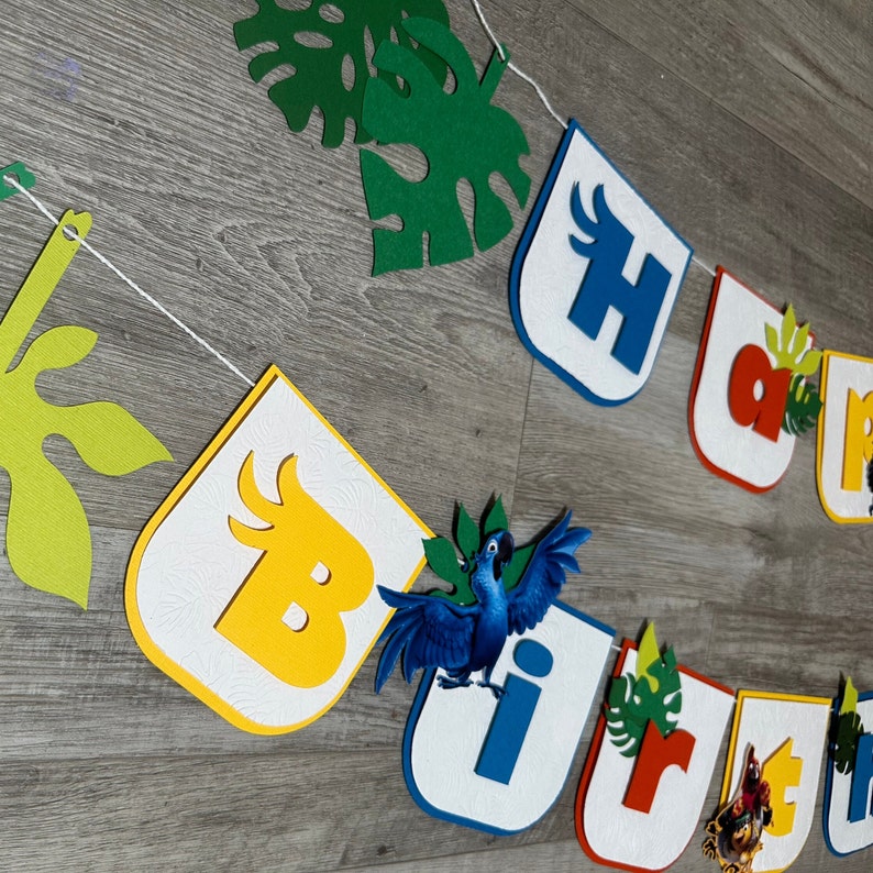 May include: A colorful felt banner with the words "Happy Birthday" in blue, red, and yellow letters. The banner also features green felt leaves and a blue bird with yellow accents.