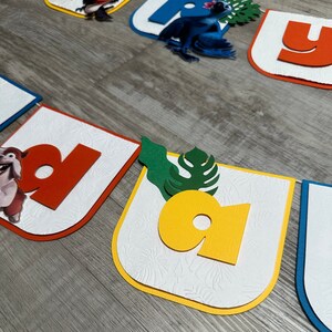 May include: A colorful birthday banner with letters that spell "Happy Birthday". The letters are cut out of red, yellow, blue, and green cardstock and are decorated with images of a parrot, a dog, and a leaf.