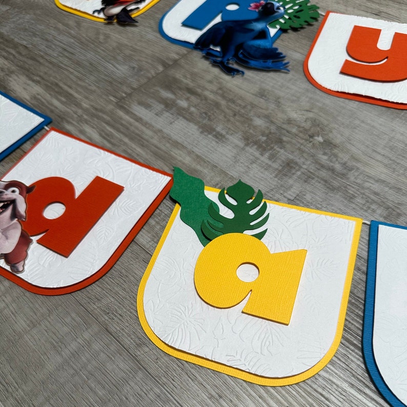 May include: A colorful birthday banner with letters that spell "Happy Birthday". The letters are cut out of red, yellow, blue, and green cardstock and are decorated with images of a parrot, a dog, and a leaf.