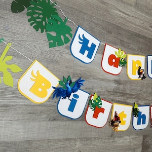 May include: A colorful felt banner with the word "Birthday" spelled out in individual letters. Each letter is decorated with a different character from the animated movie Rio, including a blue macaw, a toucan, and a yellow bird. The banner is strung together with white string and has green leaves and vines between the letters.