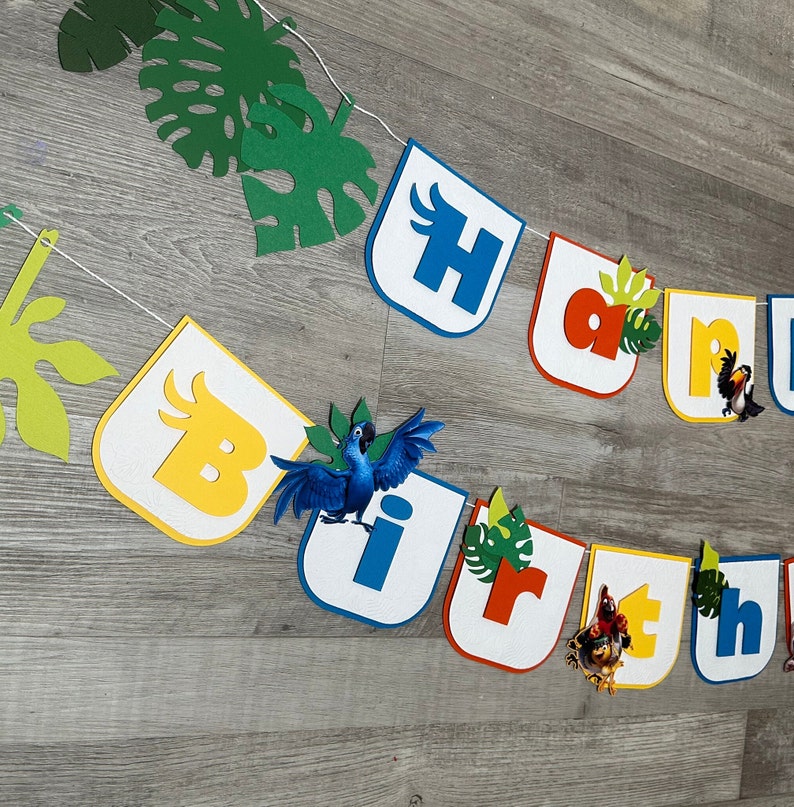 May include: A colorful felt banner with the word "Birthday" spelled out in individual letters. Each letter is decorated with a different character from the animated movie Rio, including a blue macaw, a toucan, and a yellow bird. The banner is strung together with white string and has green leaves and vines between the letters.