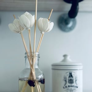 May include: A clear glass bottle diffuser with wooden sticks and white fabric flower accents. The bottle contains a dark liquid and has a label that reads "Rosebud Clean Room Diffuser Handmade Diffuser 8.5 oz".