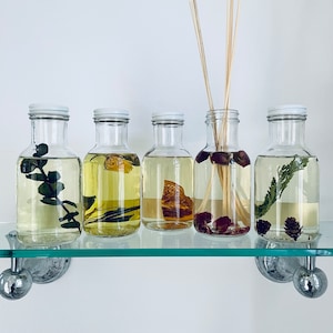 May include: Five clear glass bottles with white lids filled with different dried botanicals and liquids. The bottles are arranged on a glass shelf with silver metal brackets.