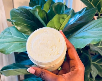 Op de afbeelding: Een witte pot met body butter met een swirl-ontwerp erop. De pot wordt vastgehouden door een hand voor een groene, bladerrijke plant.