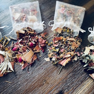 May include: Five small white mesh bags filled with dried herbs and flowers. The bags are open and the contents are spilling out onto a wooden surface.