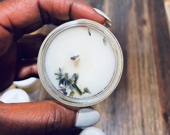Puede incluir: Una pequeña vela de vidrio con una base de cera blanca y flores de lavanda secas incrustadas en la cera. La vela está en la mano de una persona.