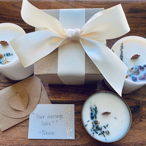 May include: Three white candles with dried lavender and crystals inside, packaged in a brown box with a white satin ribbon. A handwritten note reads "Your message here!" - Name.