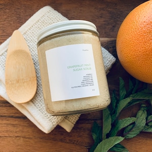 Puede incluir: Un tarro de vidrio lleno de un exfoliante de azúcar de pomelo y menta. El tarro tiene una etiqueta blanca con el texto "Grapefruit Mint Sugar Scrub" y una lista de ingredientes. El tarro está sobre una superficie de madera con una toallita blanca y un pomelo.