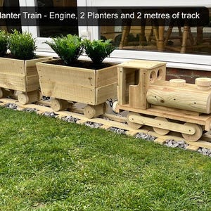 K&ouml;nnte beinhalten: Ein h&ouml;lzerner Spielzeugzug mit zwei Pflanzenwagen und einer Lokomotive. Der Zug befindet sich auf einem Gleis und hat gr&uuml;ne Pflanzen in den Pflanzenwagen. Der Text "Planter Train - Engine, 2 Planters and 2 metres of track" ist im Bild sichtbar.