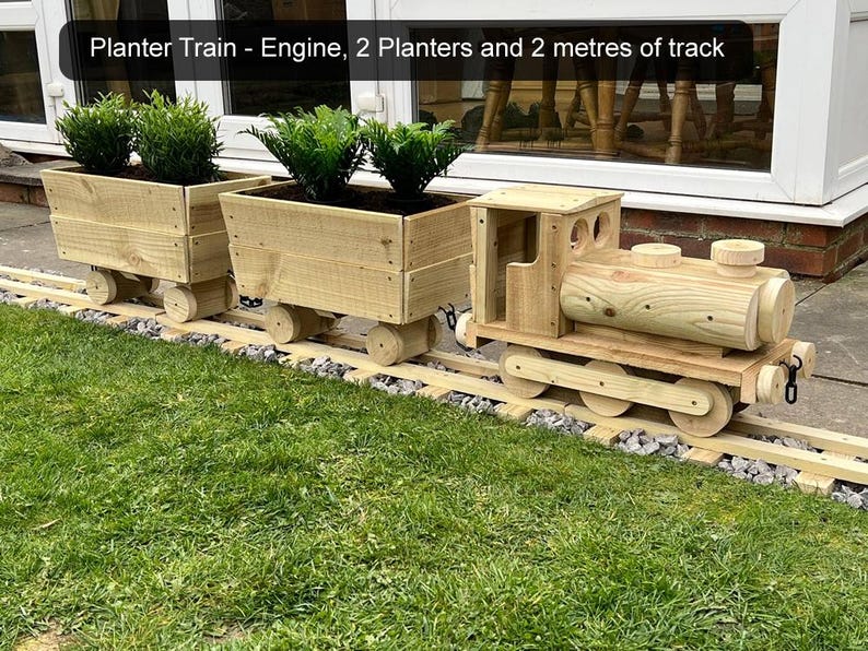 K&ouml;nnte beinhalten: Ein h&ouml;lzerner Spielzeugzug mit zwei Pflanzenwagen und einer Lokomotive. Der Zug befindet sich auf einem Gleis und hat gr&uuml;ne Pflanzen in den Pflanzenwagen. Der Text "Planter Train - Engine, 2 Planters and 2 metres of track" ist im Bild sichtbar.