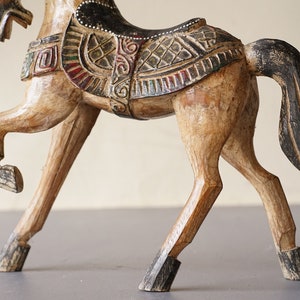 May include: A wooden horse figurine with intricate carvings and painted details. The horse is brown with black accents and has a saddle with colorful geometric patterns.