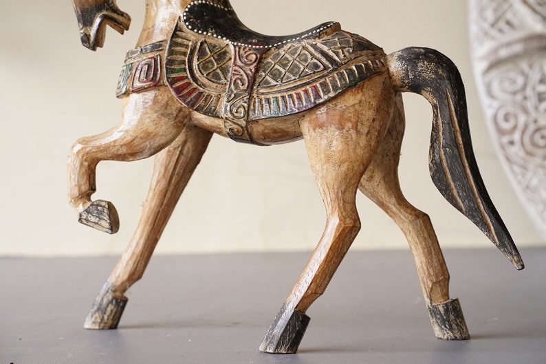 May include: A wooden horse figurine with intricate carvings and painted details. The horse is brown with black accents and has a saddle with colorful geometric patterns.