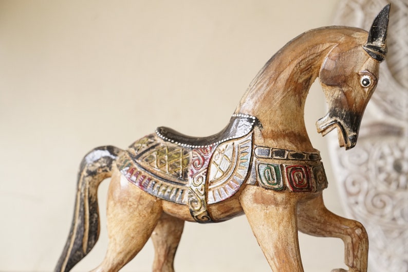May include: A wooden horse figurine with intricate carvings and colorful paint details. The horse is standing on its hind legs with its head turned to the side. The horse is decorated with a saddle and other details.
