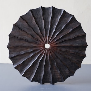 May include: A dark brown wooden wall hanging with a circular shape and a central hole. The wood is carved with radiating lines that create a sunburst pattern.