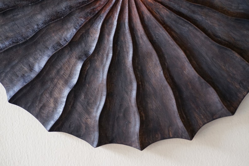 May include: A dark wood wall sculpture with a fan-like design. The wood is carved with a series of concentric circles, creating a ripple effect.