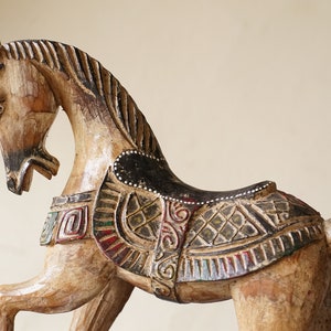 May include: A wooden horse sculpture with intricate carvings and painted details. The horse is brown with black and white accents and a saddle with a green, red, and gold design.