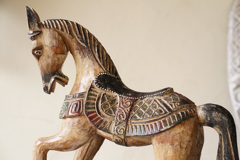 May include: A wooden horse sculpture with intricate carvings and painted details. The horse is brown with black and white accents and a saddle with a green, red, and gold design.