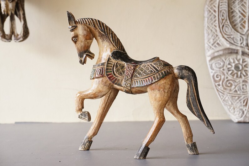 May include: A hand-carved wooden horse figurine with intricate details and colorful paint. The horse is standing on all four legs with its head turned to the side. The horse is decorated with a saddle and other embellishments.