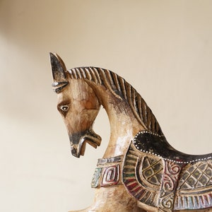 May include: A wooden horse figurine with intricate carvings and painted details. The horse is brown and white with black accents and has a saddle with colorful geometric patterns.