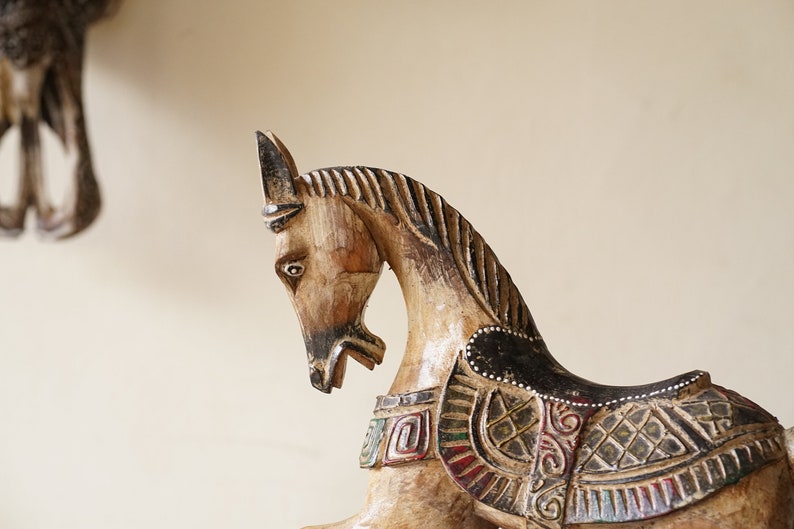 May include: A wooden horse figurine with intricate carvings and painted details. The horse is brown and white with black accents and has a saddle with colorful geometric patterns.