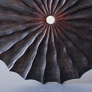 May include: A dark brown wooden wall art piece with a circular shape and a central hole. The wood is carved in a radial pattern, creating a sunburst effect.