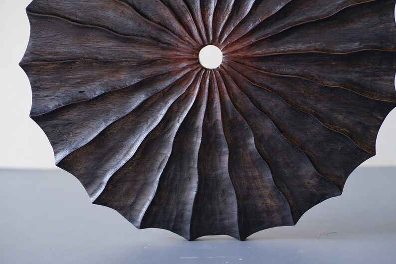 May include: A dark brown wooden wall art piece with a circular shape and a central hole. The wood is carved in a radial pattern, creating a sunburst effect.
