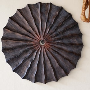 May include: A dark brown wooden wall art piece with a circular shape and a radial pattern. The center of the piece has a small hole.