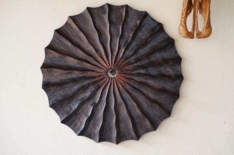 May include: A dark brown wooden wall art piece with a circular shape and a radial pattern. The center of the piece has a small hole.