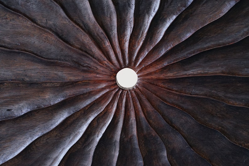May include: A close-up of a dark brown wooden surface with a circular, white, raised design in the center. The surface has a radial pattern of grooves radiating outward from the center.