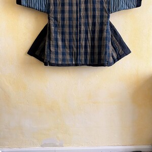 May include: A blue and brown checked quilted jacket with a striped sleeve. The jacket is hanging on a hook against a yellow wall.