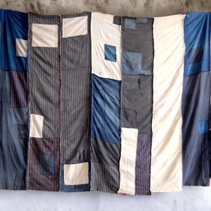 Puede incluir: Textil de patchwork colgante, con paneles verticales de diversas telas. El diseño incorpora índigo, crema y patrones de rayas. Parches de tela están cosidos a los paneles. El textil está montado en un marco de madera blanca.