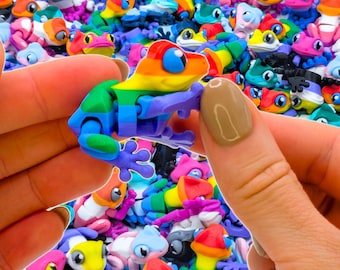 May include: A small, articulated plastic frog toy featuring a vibrant rainbow color scheme with segments in red, orange, yellow, green, blue, and purple. The frog has large, expressive eyes and a flexible body design, surrounded by a pile of similar frogs.