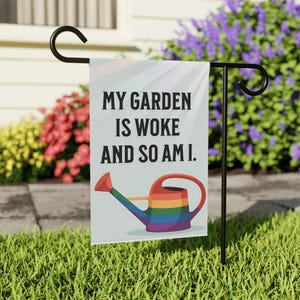 May include: A garden flag with the text "MY GARDEN IS WOKE AND SO AM I." features a rainbow-colored watering can design. The flag is displayed on a black metal stand in a garden setting with green grass and colorful flowers.