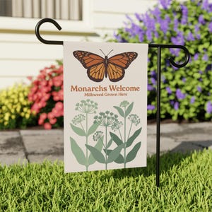 Könnte beinhalten: Eine Gartenflagge mit einer Illustration eines Monarchfalters und dem Text "Monarchs Welcome Milkweed Grown Here". Die Flagge hat einen cremefarbenen Hintergrund mit einem braunen Schmetterling und grünen Pflanzenillustrationen. Sie wird auf einem schwarzen Metallständer in einem Garten ausgestellt.