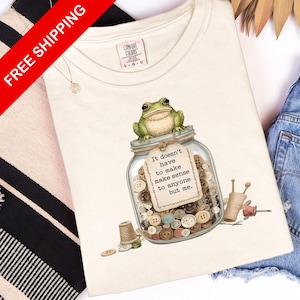 May include: Cream-colored t-shirt featuring a whimsical illustration. A green frog sits atop a jar filled with buttons, with the text "It doesn't have to make sense to anyone but me." The shirt includes a Comfort Colors tag.