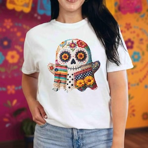 May include: White t-shirt featuring a colorful ghost design. The ghost is decorated with floral and patchwork patterns, including flowers, buttons, and polka dots. The design has a Day of the Dead theme.