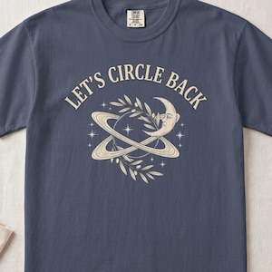 May include: A slate blue t-shirt featuring the text "LET'S CIRCLE BACK" in a cream-colored font. The graphic depicts a planet with rings, a crescent moon with a face, olive branches, and small stars in a cream-colored line art style.