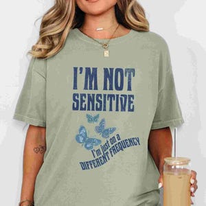 May include: Sage green t-shirt with the text "I'M NOT SENSITIVE" in dark blue block letters. Below the text are three blue butterflies and the phrase "I'm just on a DIFFERENT FREQUENCY". The shirt is a casual, oversized style.