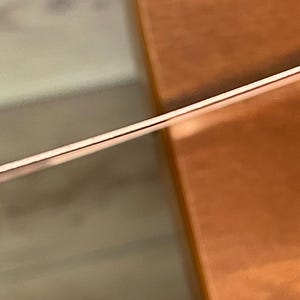 Op de afbeelding: Een dunne metalen draad met een koperkleurige afwerking, horizontaal in het kader geplaatst. De draad heeft een glad, reflecterend oppervlak dat het licht opvangt, waardoor een subtiele glans over de lengte ontstaat tegen een wazige achtergrond.