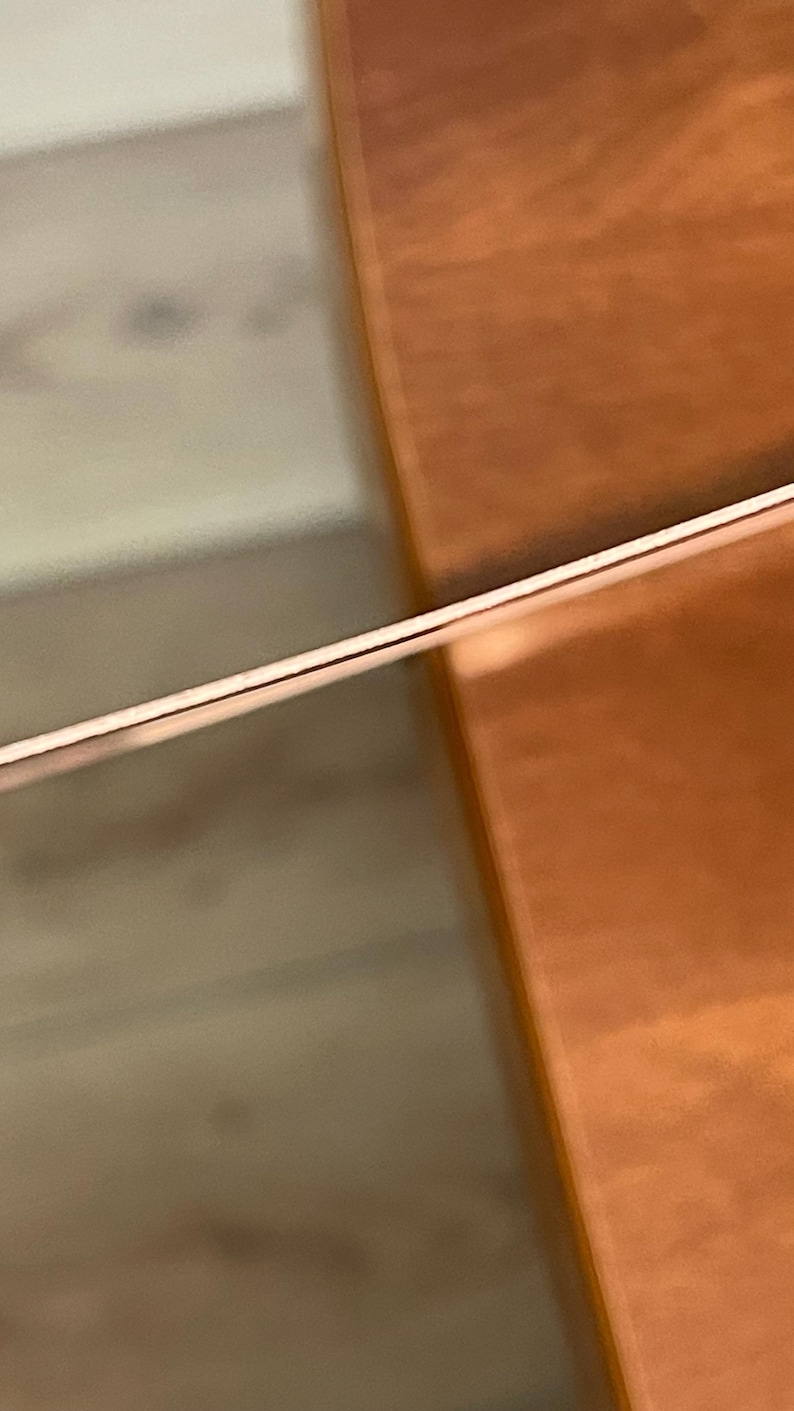 Op de afbeelding: Een dunne metalen draad met een koperkleurige afwerking, horizontaal in het kader geplaatst. De draad heeft een glad, reflecterend oppervlak dat het licht opvangt, waardoor een subtiele glans over de lengte ontstaat tegen een wazige achtergrond.