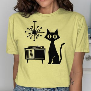 Könnte beinhalten: Hellgelbes T-Shirt mit einer schwarzen Grafik einer Katze, eines Plattenspielers und eines Kronleuchters im Mid-Century-Modern-Stil. Die Katze sitzt aufrecht. Der Plattenspieler hat eine Schallplatte.