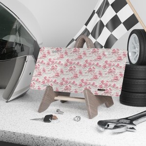 May include: A pink and white floral patterned license plate holder with a wooden stand. The license plate holder is on a table with a wrench and keys.