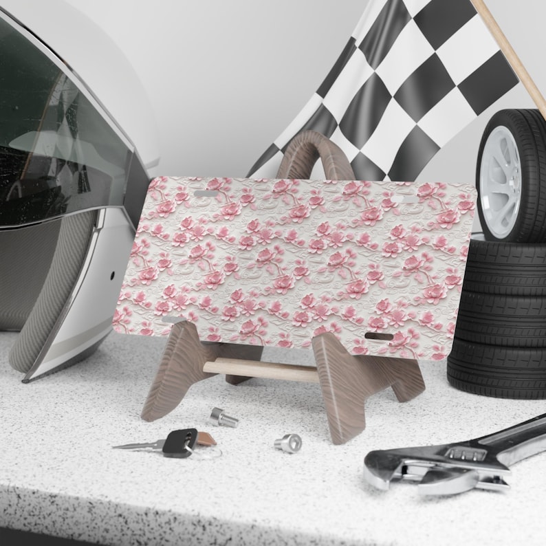May include: A pink and white floral patterned license plate holder with a wooden stand. The license plate holder is on a table with a wrench and keys.